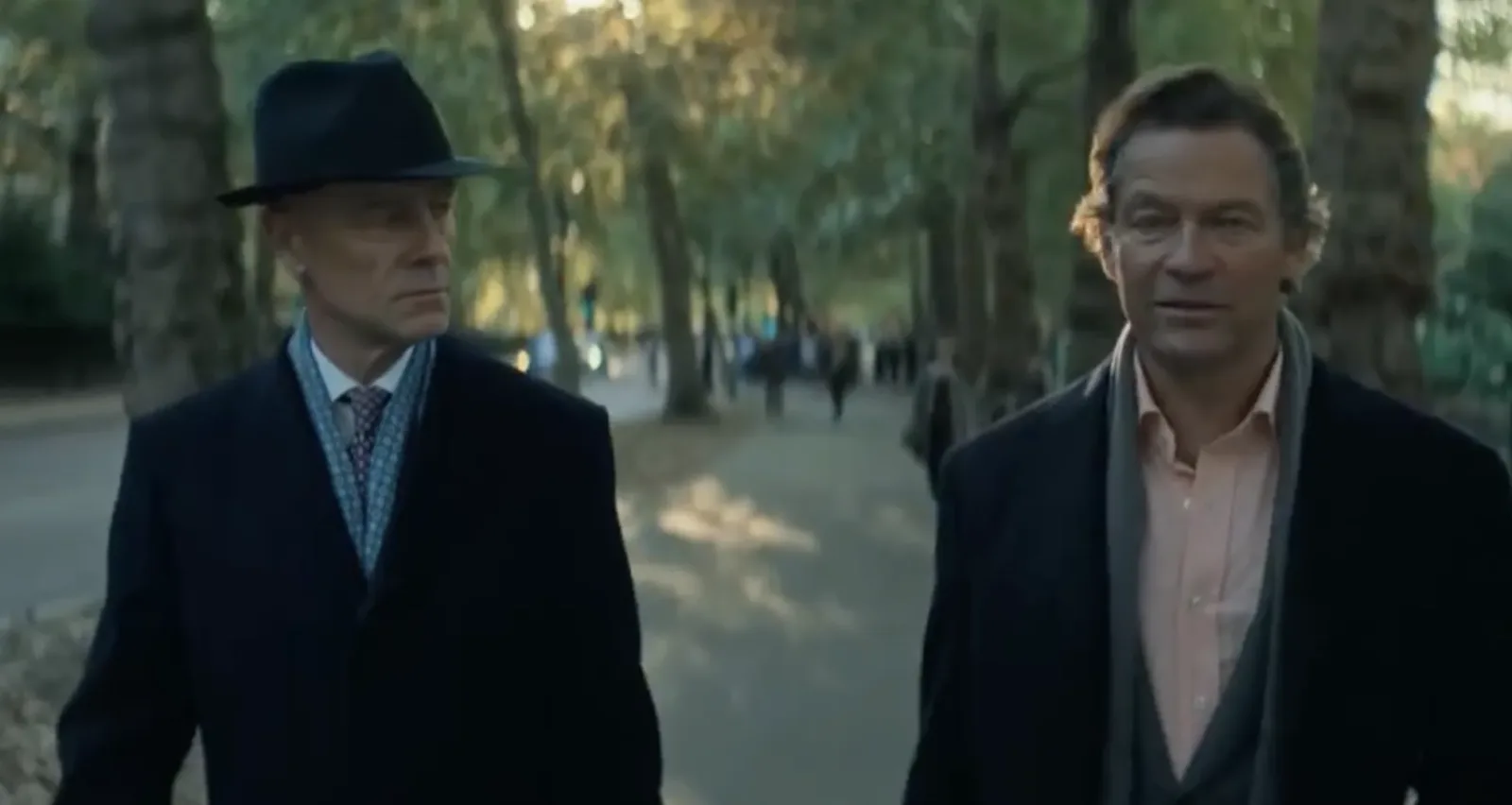 Two men in dark coats walking along a tree-lined path.